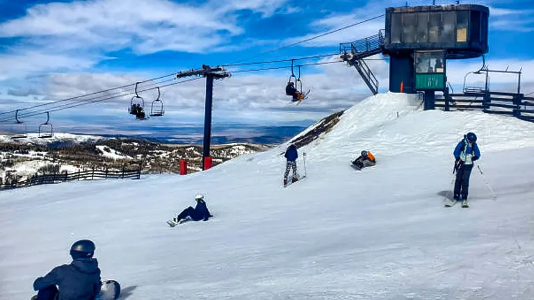 Skiers and snowboarders on a snowy hill with a ski lift overhead and a lift station in view, under a partly cloudy sky with a landscape stretching into the distance.