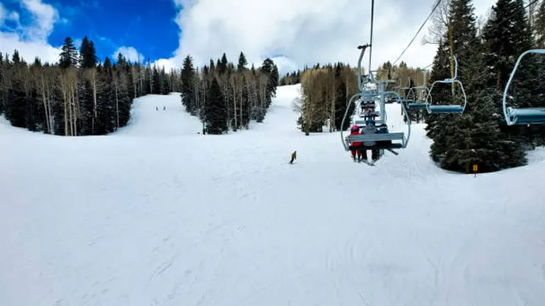 Riders ascend on a chairlift over a snow-covered ski slope dotted with trees, with a clear blue sky punctuated by fluffy clouds above.