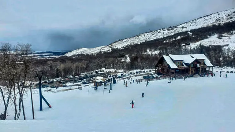 A snowy ski slope bustling with skiers near a large lodge, with ski lifts and a parking lot in view, set against a backdrop of overcast skies and rolling hills.