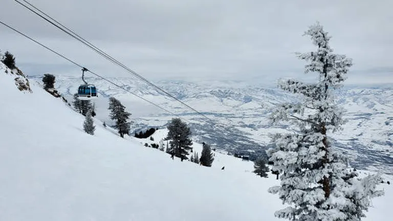 A blue ski lift cabin traveling over a snowy mountain slope with frosted trees and a view of a valley with intricate patterns of roads and plots of land in the distance, under an overcast sky.