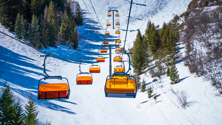 A series of bright orange ski lift gondolas move along a cable over a snowy slope lined with evergreen trees on a sunny day with shadows cast on the snow.