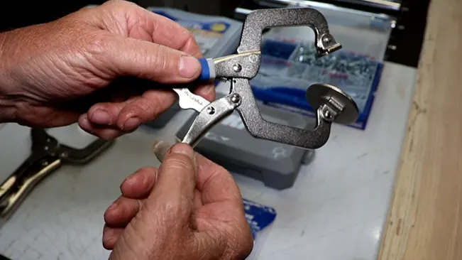 pair of hands using a metal C-clamp in a workspace, indicating some form of manual work or assembly