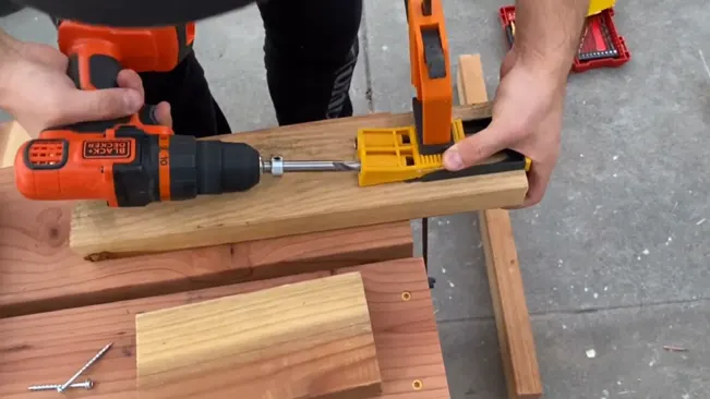 Person using an orange and black drill on a clamped piece of wood, with another piece of wood and a silver screw nearby