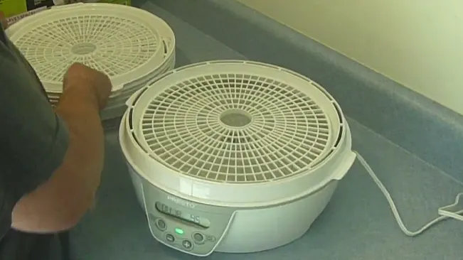 shows a hand lifting the top of a white, round, electric food dehydrator with digital controls