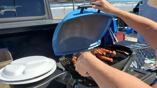 Person grilling sausages on a portable grill at the beach