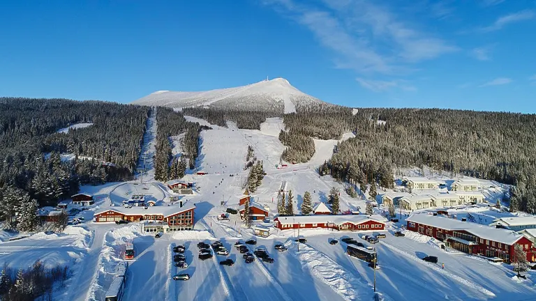 An aerial view of a ski resort with alpine buildings, parked cars, and snow-covered slopes flanked by dense forests under a clear blue sky.