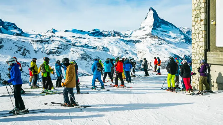A group of skiers and snowboarders gathered on a snowy slope with the iconic, pyramid-shaped Matterhorn mountain in the background under a clear sky.