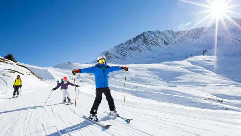 A group of skiers on a snow-covered slope with a bright sun shining in the clear blue sky and mountain peaks in the distance.