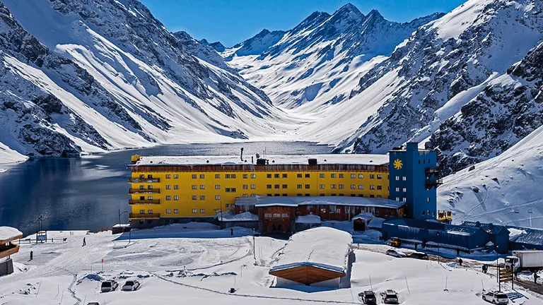 A large yellow hotel stands at the base of a snow-covered mountain valley beside a frozen lake, under a clear blue sky.