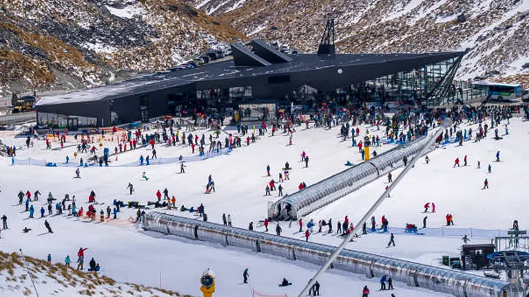 A bustling ski resort base with skiers and snowboarders near a modern lodge at the foot of a mountain slope.