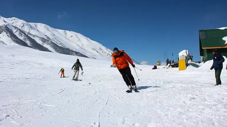 Skiers on a slope with one person in an orange jacket in the foreground, and snow-covered mountains under a clear blue sky in the background.