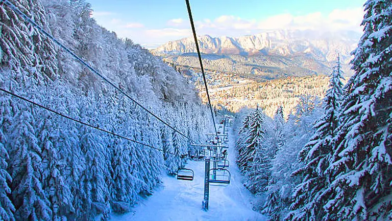 A ski lift with empty chairs ascending a snow-covered mountain, flanked by frosty trees, with a view of distant peaks under a soft blue sky.