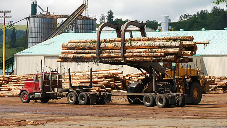 A large forklift placing a bundle of logs onto a flatbed trailer in a lumber yard, with a mill building and stacked logs in the background.
