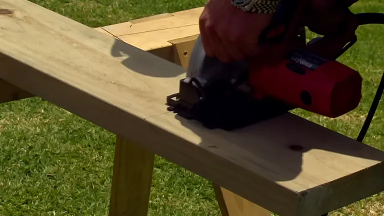 Close-up of a person using a red circular saw to cut a wooden beam on a workbench outdoors.
