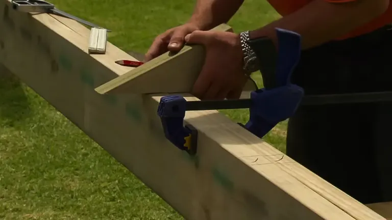 Close-up of a person using a clamp to secure a piece of wood on a beam for precise work, with tools nearby.