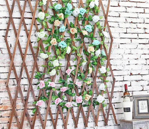 Decorative wooden trellis with an array of artificial roses in pink and green, set against a white brick wall.