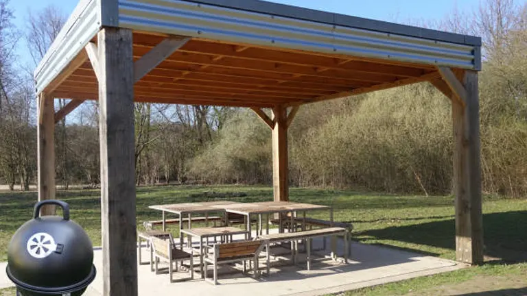 Wooden pergola with picnic tables in a park, featuring a portable grill in the foreground.
