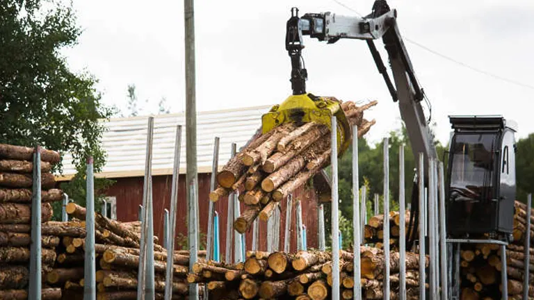 A mechanical loader is lifting a bundle of logs in a lumber yard. Stacks of cut timber are neatly arranged, ready for processing or transport. A red building is visible in the background, along with some trees, indicating the yard's rural or semi-rural setting. The scene highlights industrial logging activities and the handling of harvested timber.
