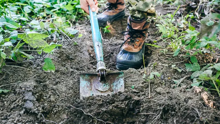 Person using a garden hoe to prepare soil for planting, wearing camouflage pants and sturdy boots.
