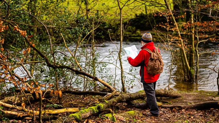 A person wearing a red jacket and a beanie stands by a forest river, holding a map. They have a brown backpack and are surrounded by trees and fallen branches, exploring the natural environment.
