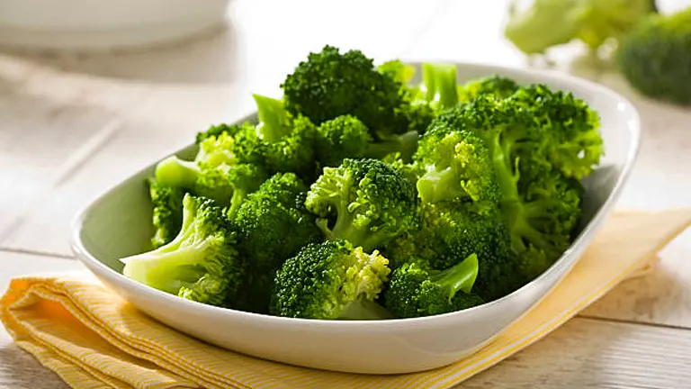 A bowl of freshly steamed broccoli florets on a white dish, placed on a yellow striped napkin on a wooden table, highlighting the vibrant green color and healthy appearance of the broccoli.