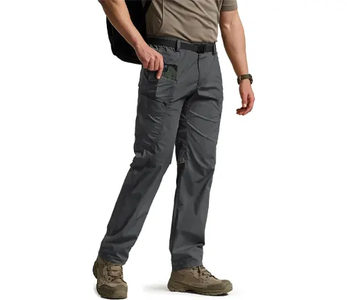 A man walking in gray hiking pants with multiple pockets, paired with a light brown t-shirt and beige hiking boots, carrying a black backpack.