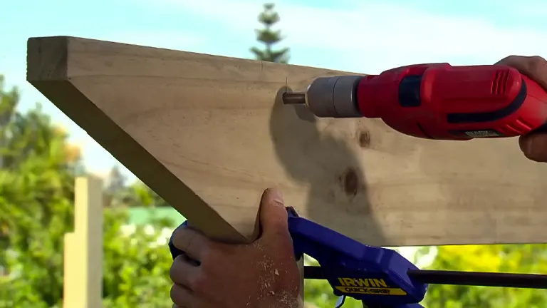 Close-up of a person using a red power drill to drive a screw into a wooden beam clamped for stability.