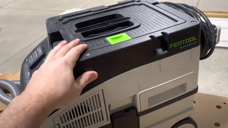 A close-up of a hand resting on the top of a Festool CT 15 E Dust Extractor, showing its black handle, green latch, and detailed control panel on a gray body