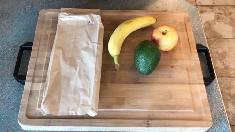 Avocado sitting on top of a chopping board with a banana, an apple, and a paper bag.