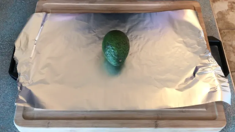 Green avocado sitting on top of foil, with a chopping board underneath.