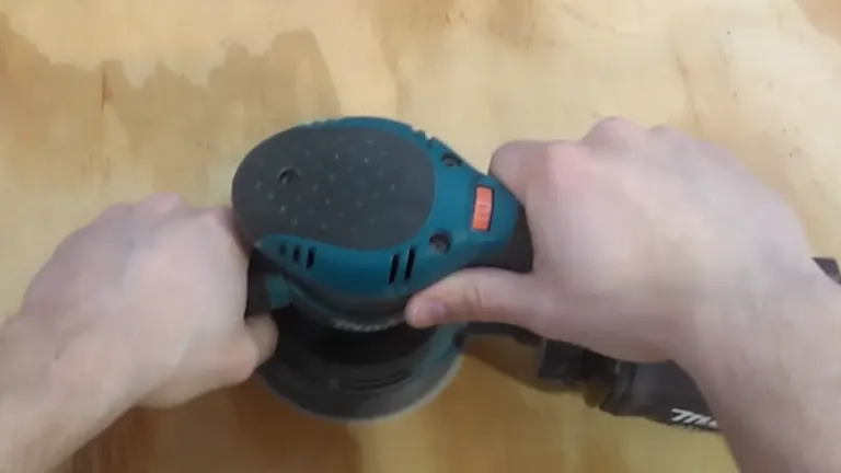 Close-up of hands operating a blue and black orbital sander on wood.