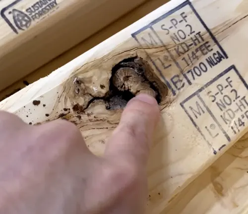 A finger pointing at a large knot and defect in a piece of wood, highlighting an area of concern for structural integrity.