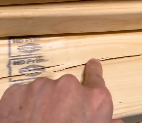  A finger pointing at a significant crack in a piece of wood, showing a defect that could compromise the wood's stability.