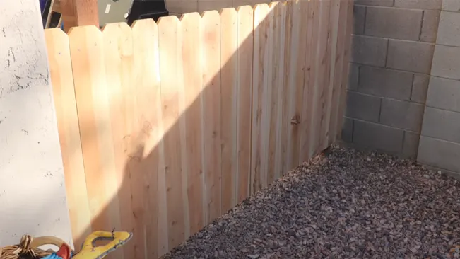 New wooden fence in a corner with a shadow cast across, adjacent to a concrete wall.
