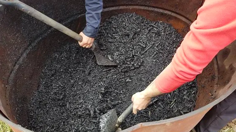 Two people using shovels to mix biochar in a large metal container outdoors.