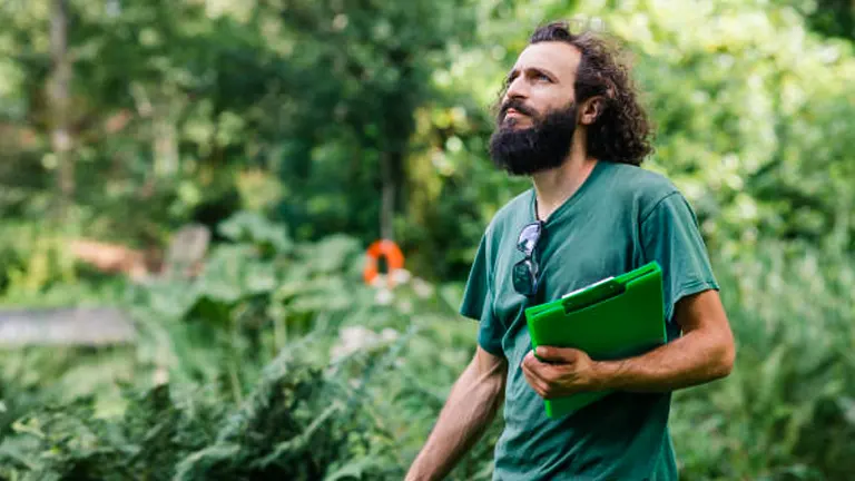 A man with a long beard and curly hair, wearing a green t-shirt, holding a clipboard and looking thoughtfully at a garden.