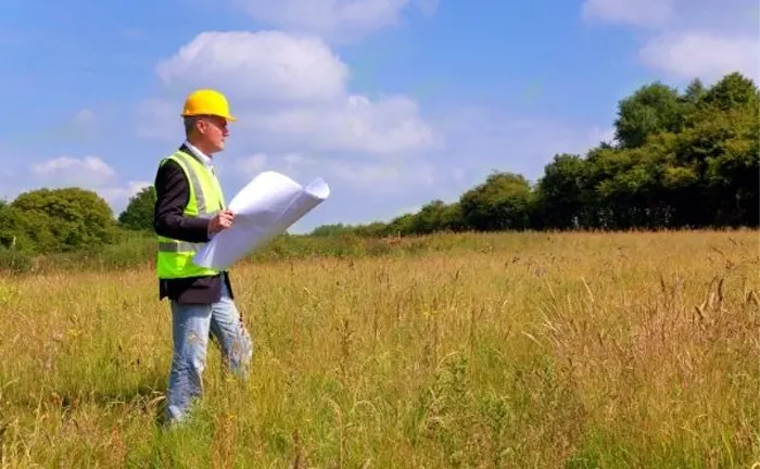 Person in a hard hat and safety vest holding blueprints on open land, assessing for sustainable forestry practices