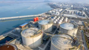 Aerial view of a large industrial oil and gas storage facility near the water, with multiple storage tanks and pipelines, surrounded by other industrial infrastructure and distant mountains in the background.