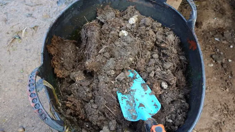 A blue plastic garden tool resting on a pile of coarse compost in a large black bucket, highlighting soil preparation activities.
