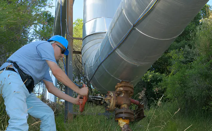 Worker adjusting a valve on industrial pipes in a forest area.