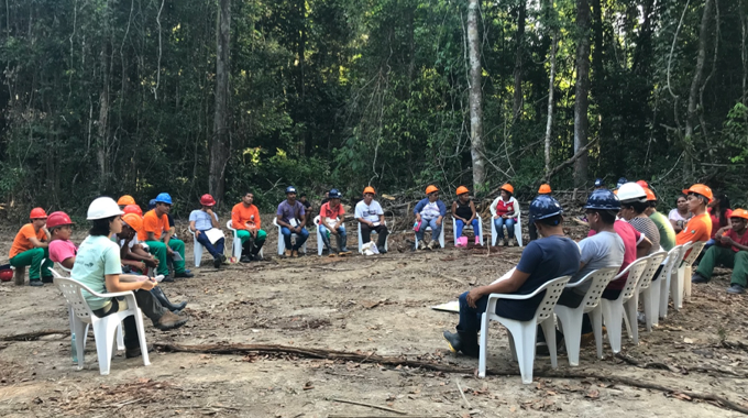 A group of workers wearing hard hats and safety gear sit in a circle on plastic chairs in a forest clearing, participating in an outdoor training session or meeting.