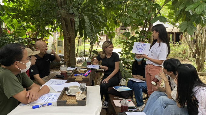 A group of people gathered outdoors in a forest setting, participating in a discussion or workshop. 