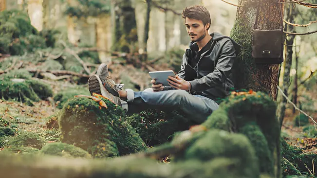 A young man sitting on mossy ground in a forest, using a tablet, representing the exploration and study involved in a Forestry and Arboriculture Career.