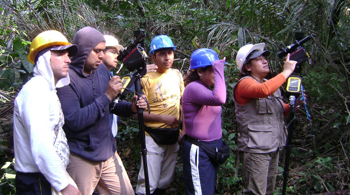 A group of people in a forest setting, wearing hard hats and outdoor gear, are using various pieces of equipment, including cameras and GPS devices.