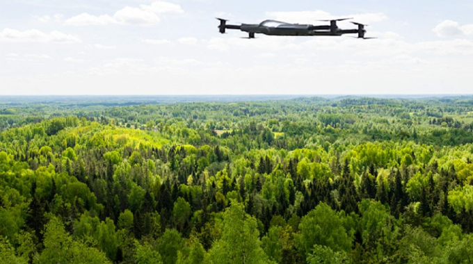 A drone flying over a vast, dense forest with a mix of deciduous and coniferous trees. The forest extends to the horizon under a partly cloudy sky,