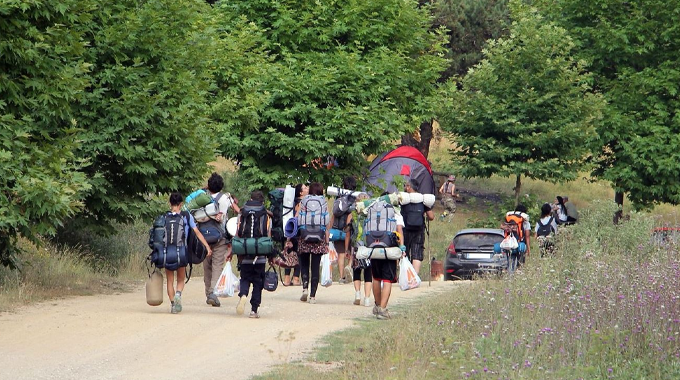 A group of hikers with large backpacks and camping gear walking along a dirt path surrounded by greenery. 