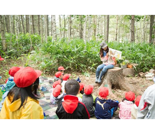 A group of young children wearing red caps sits on the ground in a forest, attentively listening to a woman reading a book while sitting on a tree stump, illustrating an outdoor educational activity promoting environmental awareness and learning.