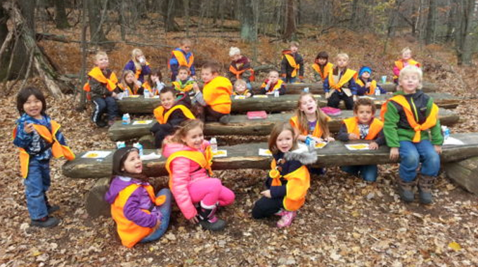 A group of children wearing orange safety vests are seated on log benches in an outdoor forest setting, participating in an educational activity.