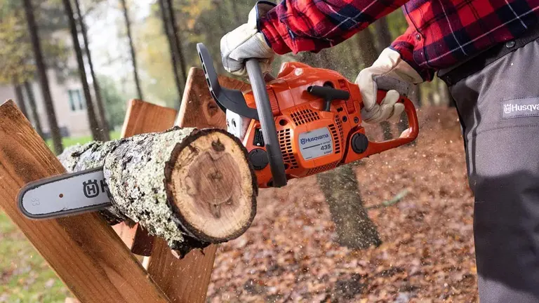 Individual operating a Husqvarna chainsaw to saw through a log held in a sawhorse.
