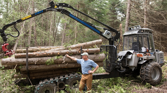 A man is standing next to a logging machine equipped with a mechanical arm, which is loading cut logs onto a trailer in a forested area.